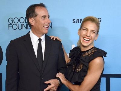 Jessica Seinfeld is grabbing Jerry Seinfeld as she is laughing, whereas, Jerry is looking somewhere.